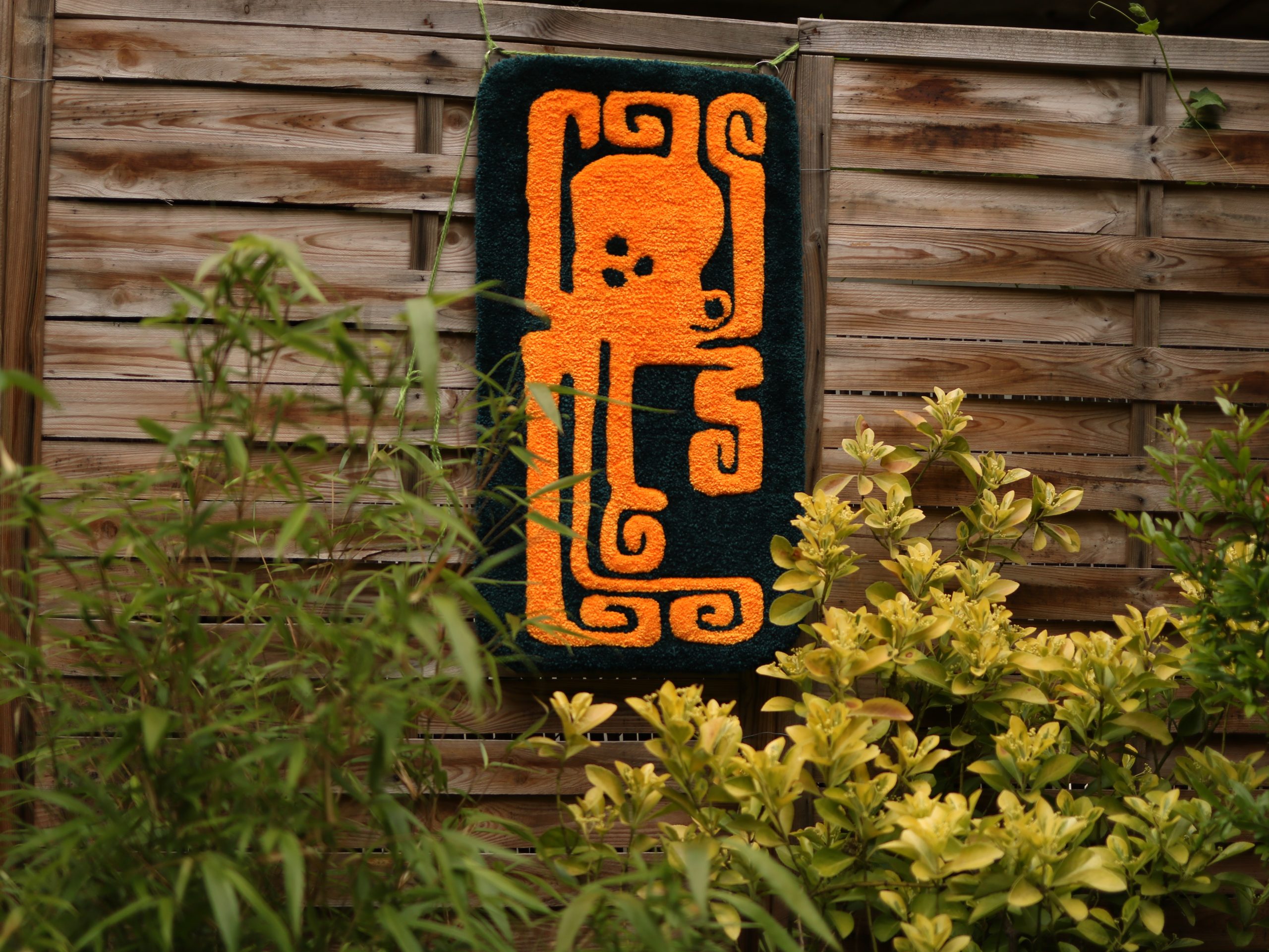 photo d'un tapis tufté avec un motif de poulpe orange, accroché à une palissade derrière des feuillages